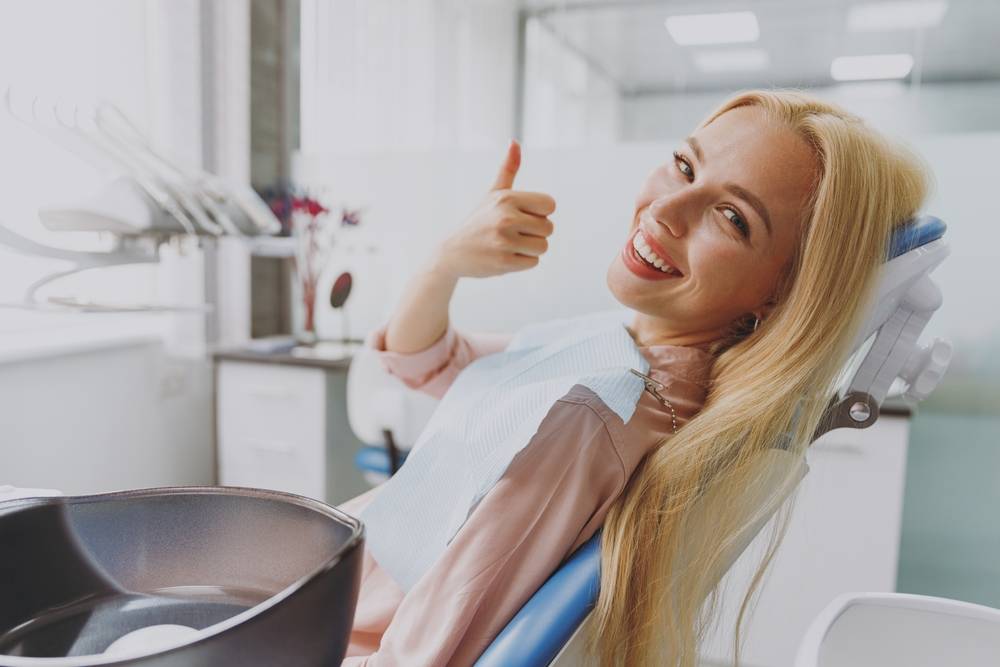 Close up young smiling happy woman 20s covered by napkin show thumb up gesture sit at dental office chair indoor cabinet waiting stomatologist for oral procedure Healthcare caries enamel treatment.
